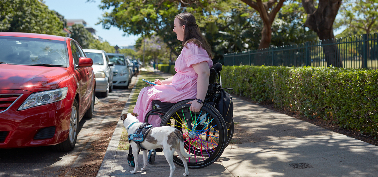 A woman in a light pink dress sits in a wheelchair with rainbow spoked wheels. Her certified assistance dog is beside her and she is holding her phone. She is on the footpath, facing a red car.